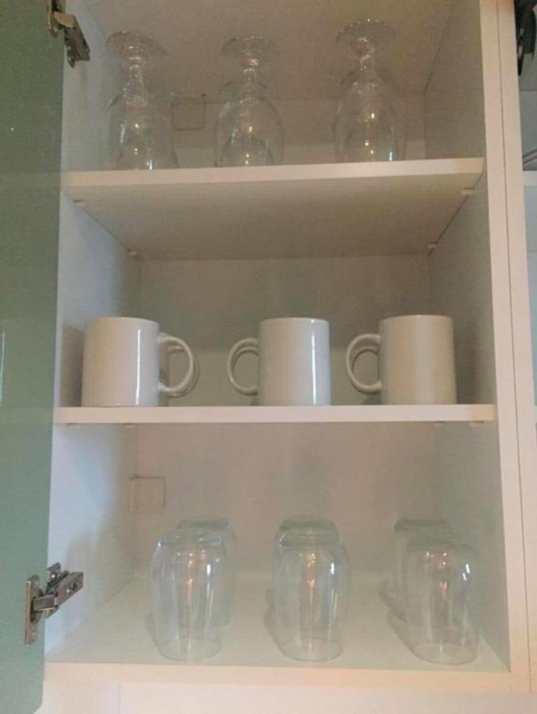 Un meuble avec trois étagères : l’étagère supérieure contient trois bouteilles en verre, l’étagère du milieu abrite trois tasses blanches, et l’étagère inférieure contient trois tasses en verre transparent.