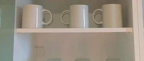 A cabinet with three shelves; the top shelf holds three glass bottles, the middle shelf has three white mugs, and the bottom shelf contains three clear glass mugs.