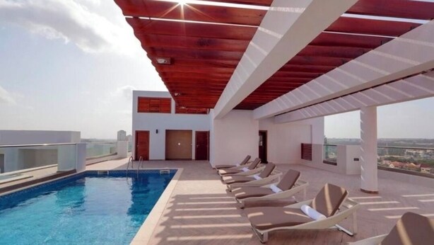 A rooftop pool area with a clear view of the city skyline, a wooden ceiling, and sunlit lounge chairs.