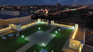 A rooftop terrace with a view of the city at night, featuring a grassy area, tables, and chairs.