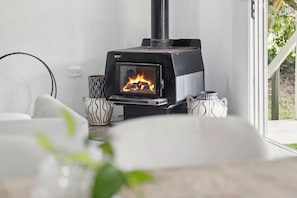 A modern living room with a wood-burning stove, a sofa, and a window with a view of greenery. - Magnolia Cottage (Leura)