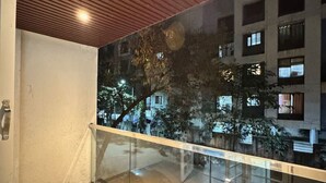 A balcony with a glass railing overlooking a building with multiple windows and trees. - Canary Haven: 2BHK Modern AC Flat in Pimple Nilakh (Pimpri-Chinchwad)