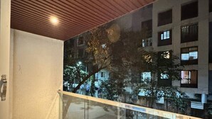 A balcony with a glass railing, wooden ceiling, and a view of a building with multiple windows. - Seagull Nest: AC 2BHK Apartment by Bleisure (Pimpri-Chinchwad)