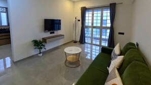 A modern living room with a green sofa, a flat-screen TV, a round coffee table, and a large window with curtains. - Crane House: Spacious 2BHK AC Flat by Bleisure (Pimpri-Chinchwad)