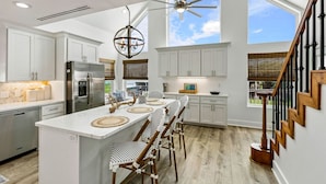 A modern kitchen with white cabinets, a central island with bar stools, a stainless steel refrigerator, and a ceiling fan. - Cozy Coastal Home, Minutes from the Beach (Jamaica Beach)