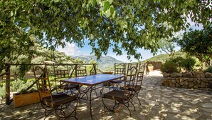 Une terrasse extérieure avec une table et des chaises, entourée de murs en pierre et de verdure, offrant une vue sur un paysage montagneux.