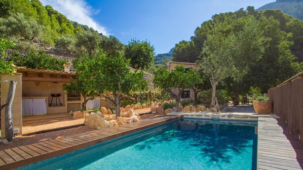 Une piscine dotée d’une terrasse en bois, entourée de rochers et d’arbres, avec des bâtiments visibles au loin.