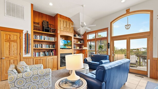Un salón amplio con una televisión grande, estanterías de madera, un sofá azul y vistas al exterior.