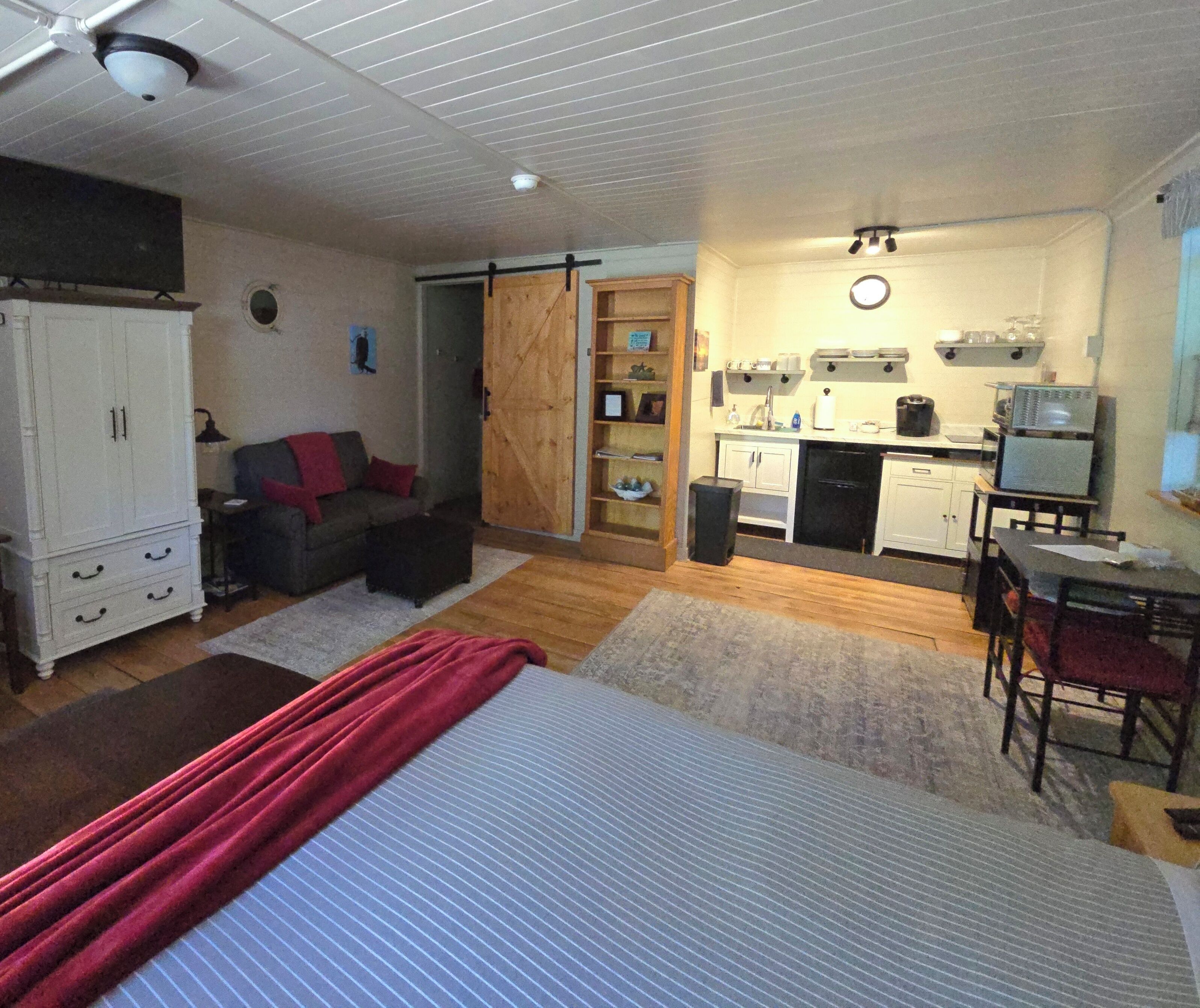 A compact living space with a kitchenette, a bed with a red blanket, a sofa, and a wardrobe.