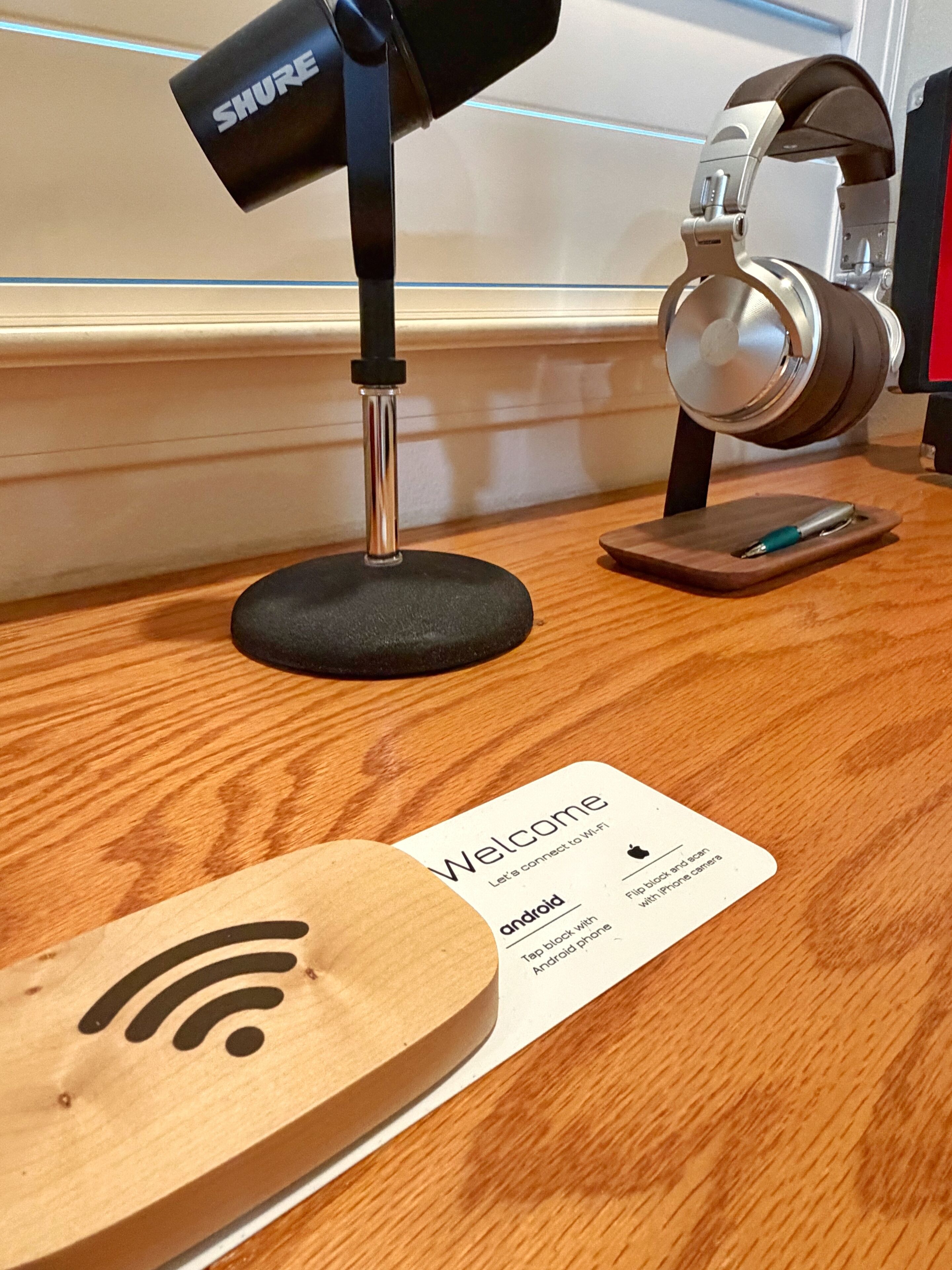 Uma escrivaninha de madeira com um microfone, fones de ouvido, um smartphone e um cartão com os dizeres 'Welcome' e 'android'.