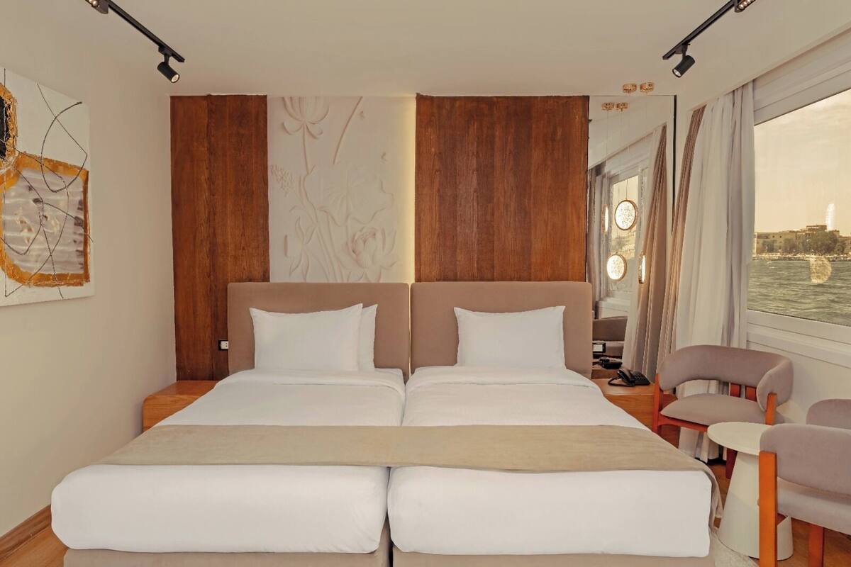 A double bed with two pillows, a wooden headboard, a bedside table with a lamp, and a view of the water.