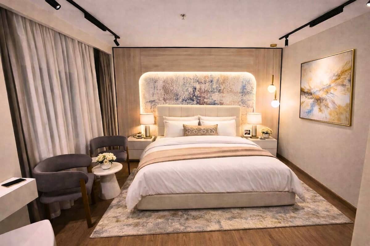 A modern hotel room with a large bed, a bedside table with a lamp, a chair, and a small table with flowers.