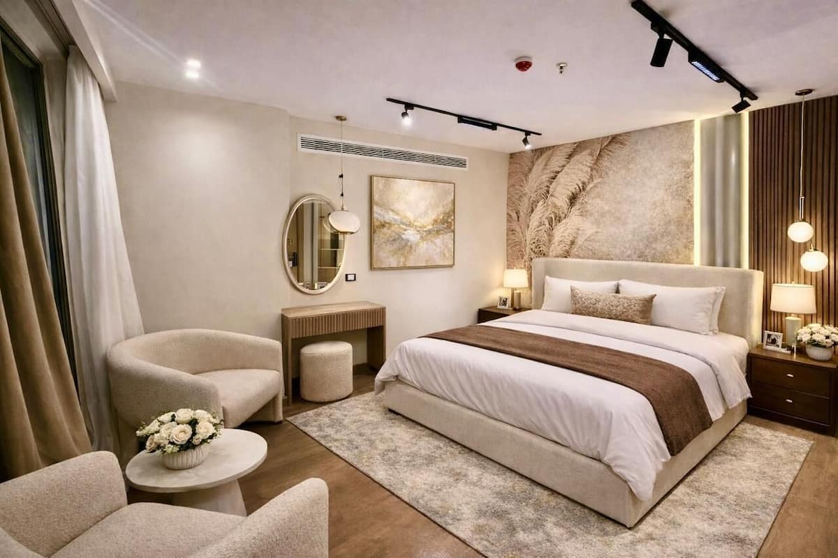 A modern bedroom with a large bed, a sitting area with a chair and a small table, a bedside table with a lamp, and a decorative mirror.