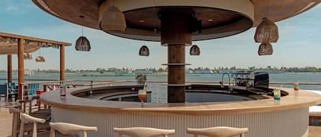 Un espace bar avec des chaises en osier, un bar circulaire et une vue sur l’eau.