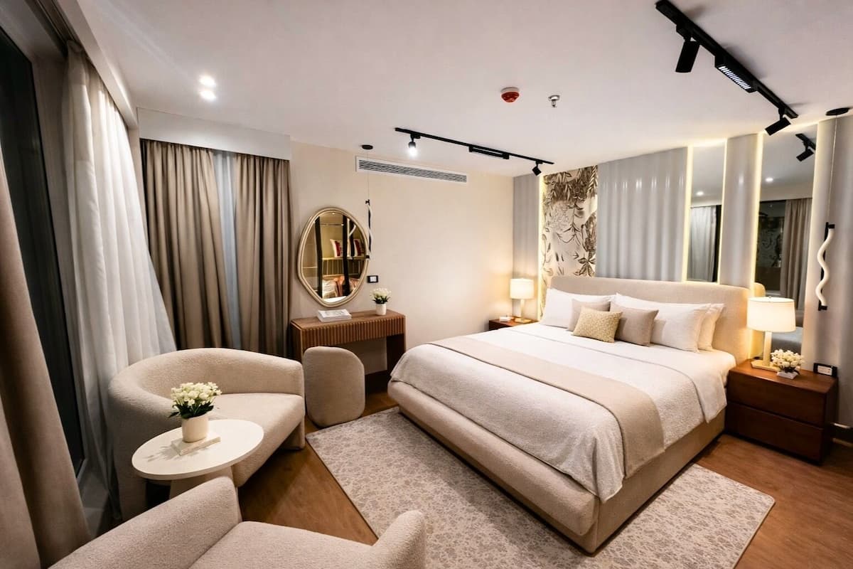 A hotel room with a large bed, two armchairs, a small table with a vase of flowers, a bedside table with a lamp, and a large mirror on the wall.