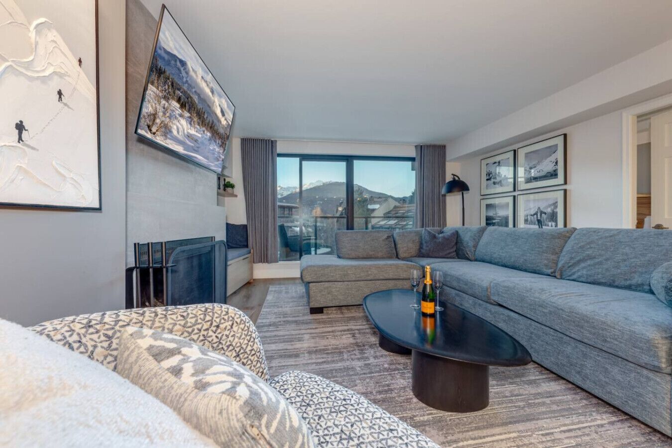 Un salon moderne avec un canapé gris, un pouf à motifs et une table basse noire sur laquelle se trouvent une bouteille et des verres.