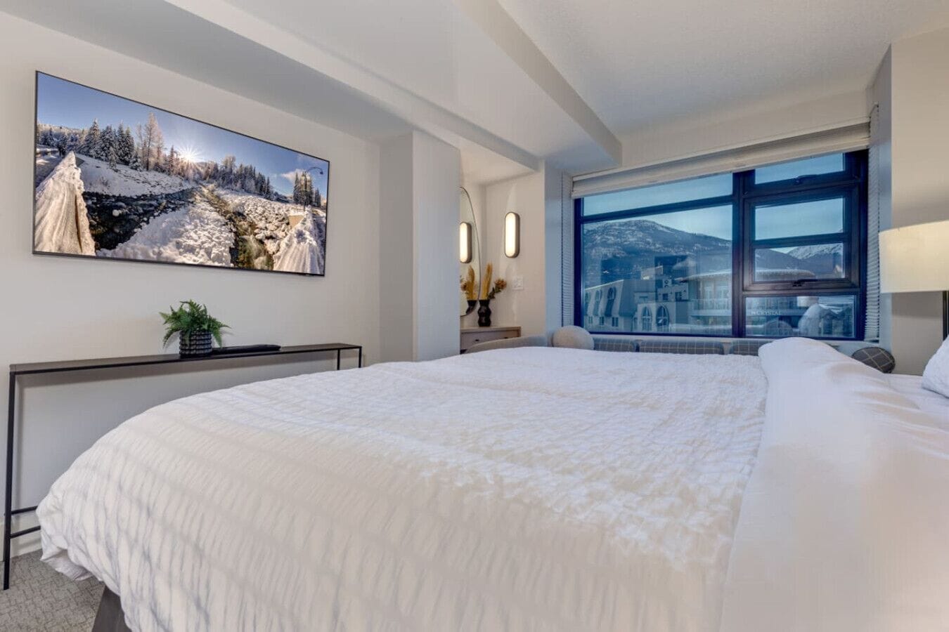 A modern hotel room with a large bed, a flat-screen TV mounted on the wall, a bedside table with a lamp, and a large window offering a view of a mountainous landscape.