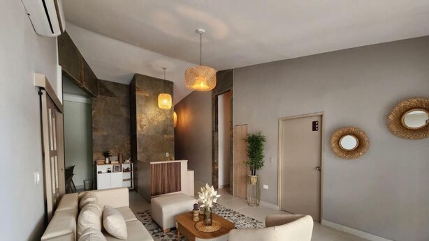 Ein modernes Wohnzimmer mit einer Couch, Sesseln, einem Couchtisch und einem Essbereich. Im Hintergrund befindet sich eine Küche, eine Tür sowie dekorative Spiegel an der Wand.