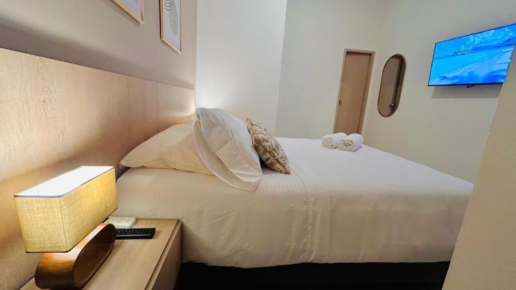Una habitació d'hotel amb un llit, una taula de nit amb una làmpada, un comandament a distància, un televisor fixat a la paret i una porta.