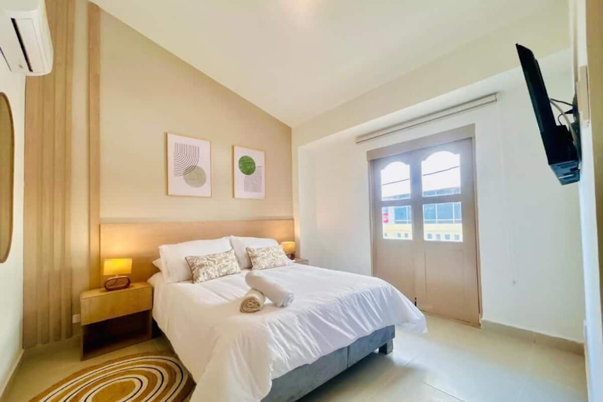 A hotel room with a bed, a nightstand, a wall-mounted TV, and a window with a view of buildings.