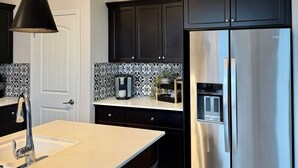 Una cocina moderna con armarios oscuros, un refrigerador de acero inoxidable, una lámpara colgante y un salpicadero de azulejos.