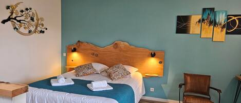 Une chambre d’hôtel avec une tête de lit en bois, un lit avec du linge de maison blanc et une couette turquoise, une table de chevet en bois, une chaise et des œuvres d’art murales.
