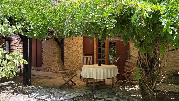 A stone patio with a round table and four wooden chairs, surrounded by lush greenery and a rustic stone wall. - Le Tuquet de Saint Chamassy, private swimming pool (ST CHAMASSY)