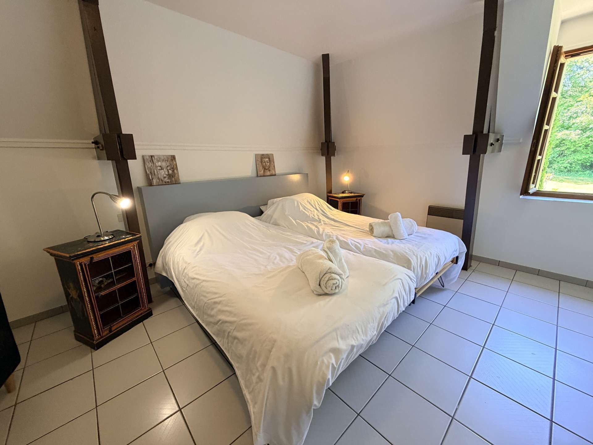 Un dormitorio con cama, mesitas de noche, una lámpara y una ventana.