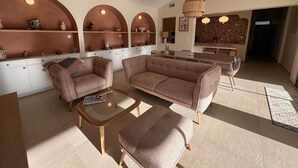 A modern living room with a sofa, armchairs, and a coffee table. There are shelves with decorative items and a kitchen area in the background. - Ref 038 - Exceptional villa with private pool and jacuzzi in the center of Soulac-sur-Mer (SOULAC SUR MER)