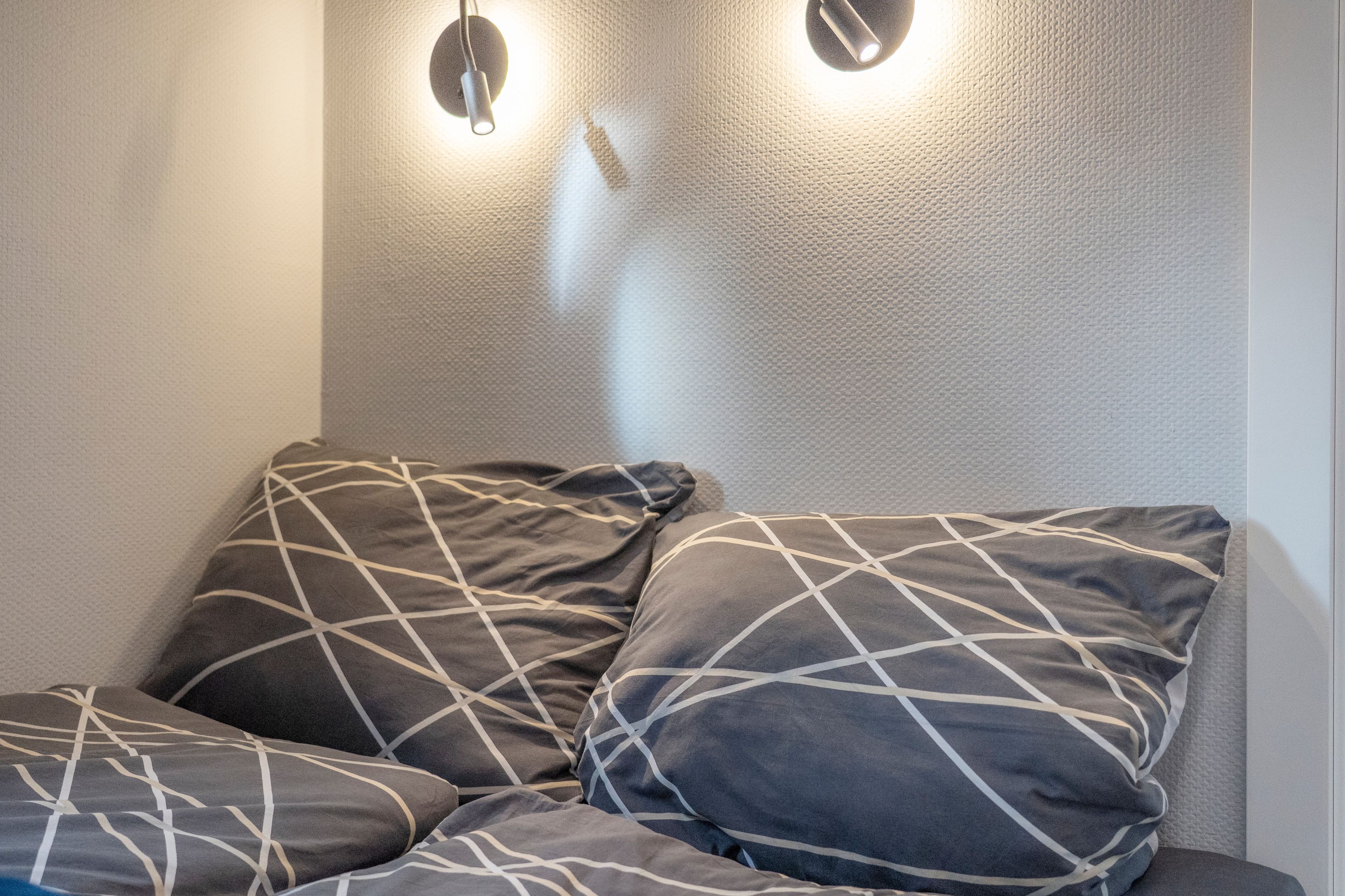 Ein Raumeck mit einer strukturierten Wand und einer Leuchte. Auf einem Sofa liegen zwei Kissen mit einem geometrischen Muster.