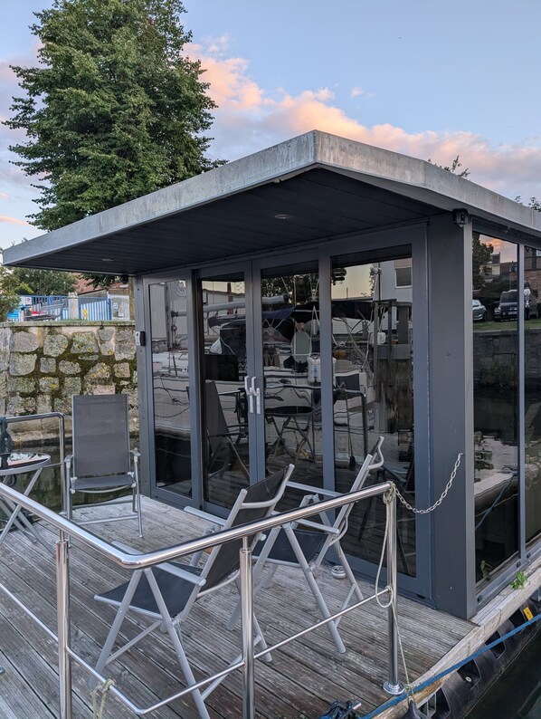 A covered outdoor seating area with chairs and tables, a stone wall, and a view of the water.