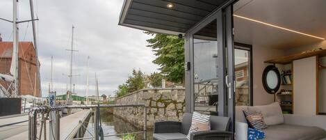 Una terraza moderna al aire libre con vistas al puerto deportivo, un ventanal que da a una sala de estar con sofá y cojines, y una pequeña mesa con una planta en maceta.