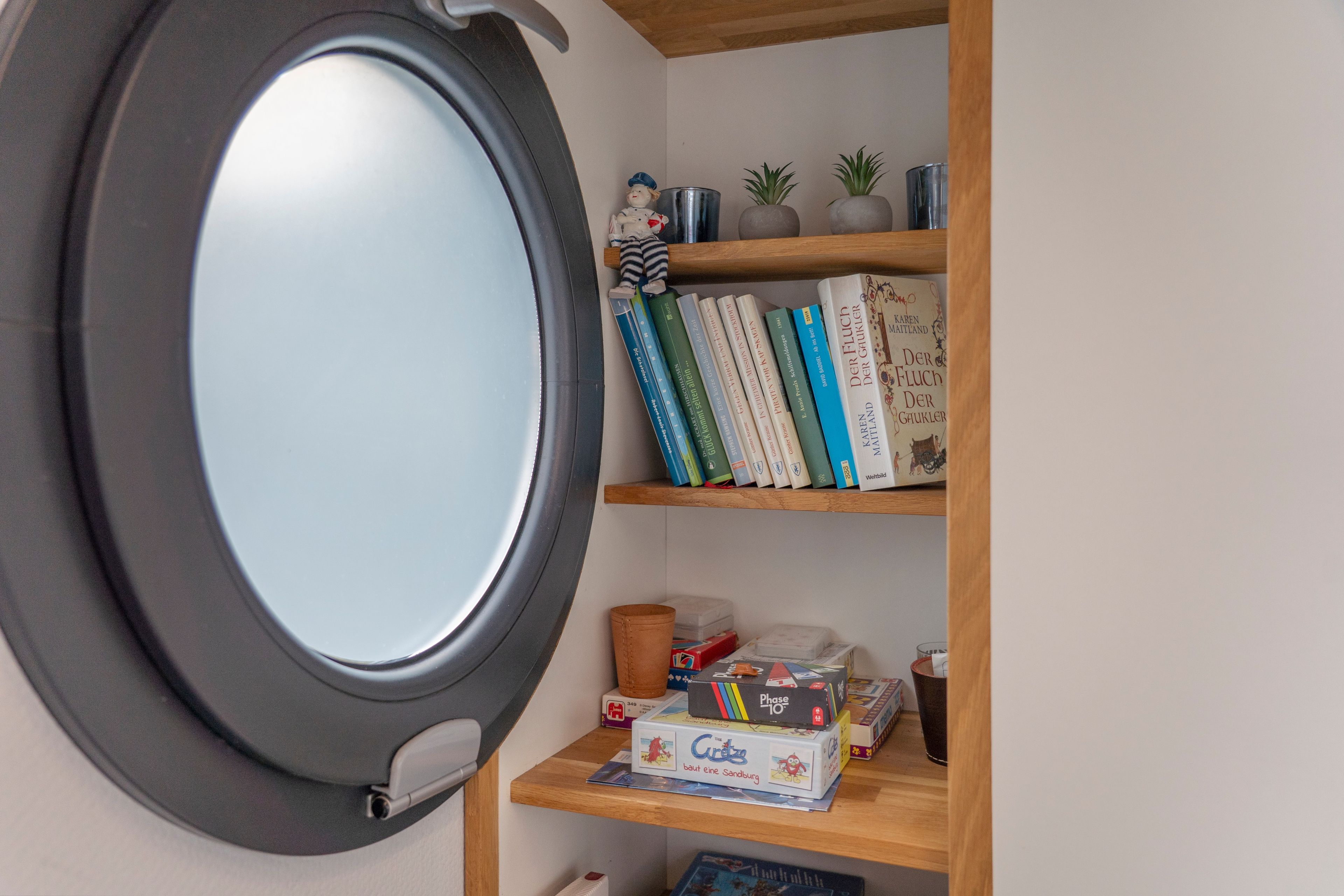 Een kamer met een rond raam, een boekenkast met boeken en decoratieve objecten, en een kleine tafel met een spel.