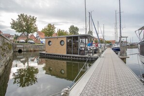 Ein schmaler Wasserweg mit einem hölzernen Steg, der zu einem kleinen Gebäude mit großen Fenstern führt.