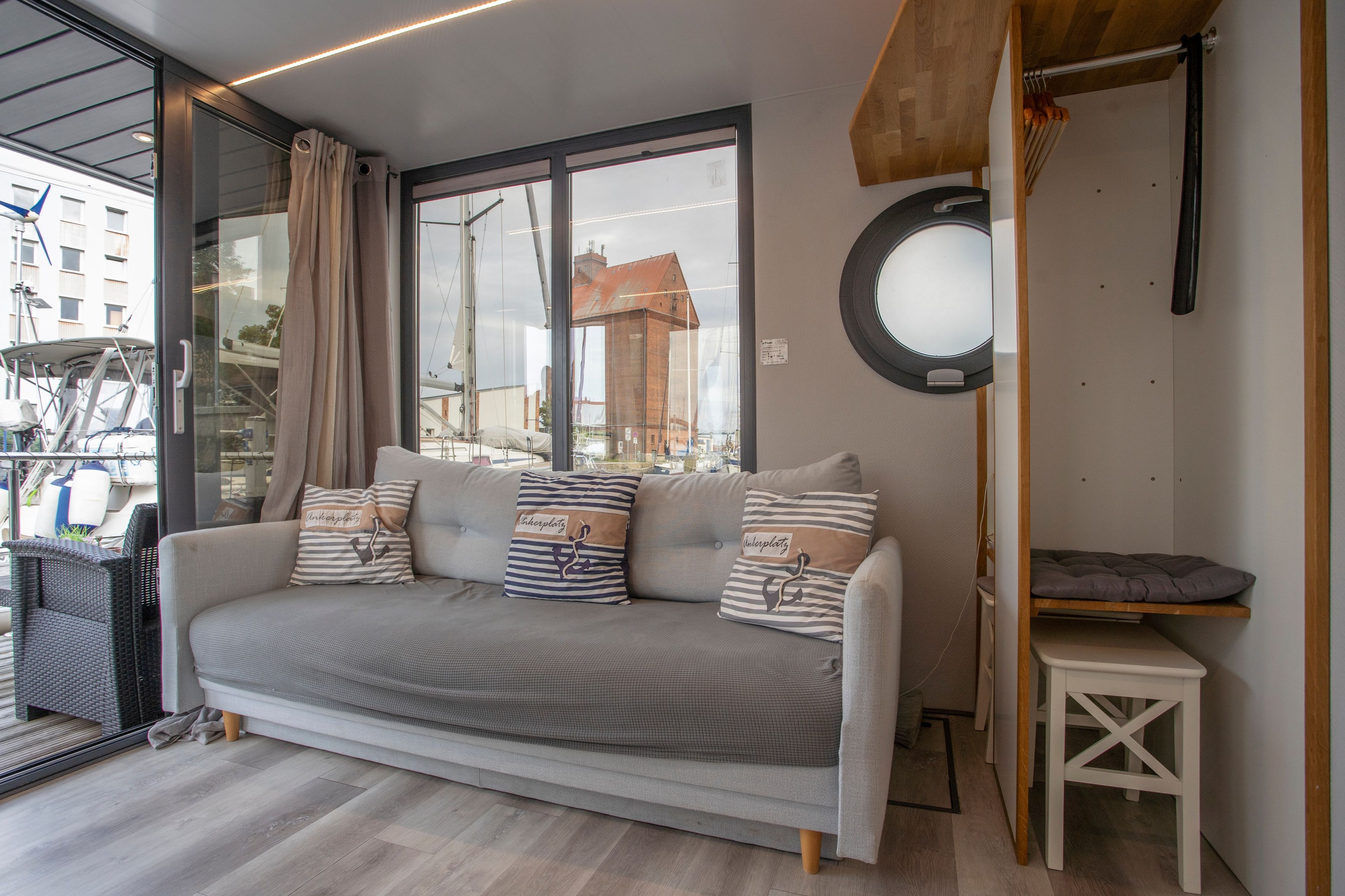 Een moderne woonkamer met een grijze bank, gestreepte kussens, een klein wit bijzettafeltje en een uitzicht op het exterieur door grote ramen.