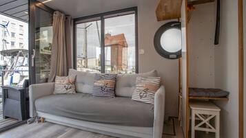Een moderne woonkamer met een grijze bank, gestreepte kussens, een klein wit bijzettafeltje en een uitzicht op het exterieur door grote ramen.