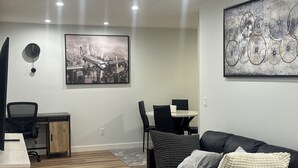 Un salon moderne avec un canapé en cuir noir, une table basse ronde, un bureau avec une chaise et un coin repas avec une table et des chaises. Les murs sont ornés d’une grande œuvre d’art encadrée représentant un paysage urbain et des bicyclettes.