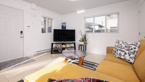 Ein Wohnzimmer mit gelbem Sofa, Glastisch, Flachbildfernseher und einem kleinen Beistelltisch mit Dekoration.