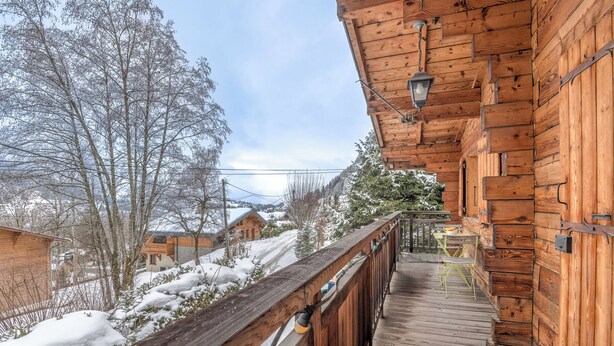 A wooden deck with a railing, a table, and chairs, overlooking a snowy landscape with houses and trees. - Chalet du Grand Air - Welkeys (Megève)
