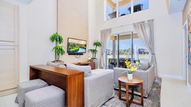 A modern living room with a sofa, a coffee table, and two ottomans. There is a TV mounted on the wall, a plant, and a window with curtains. - Beautiful Magic Village Townhome Minutes from Disney (Kissimmee)