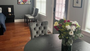 En moderne stue med mørkt trægulv, en grå sofa med knapper, et spisebord med en vase med blomster og et stort vindue med persienner.