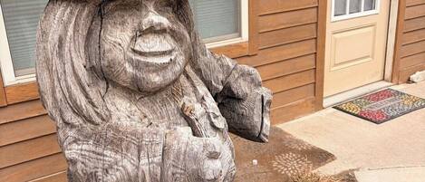 A stone statue of a figure with a hat and a pipe, placed in front of a house with a door and a wreath on the wall.
