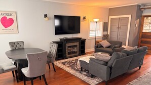 En stue med pejs, et fladskærms-tv, et spisebord med stole og en grå sofa.