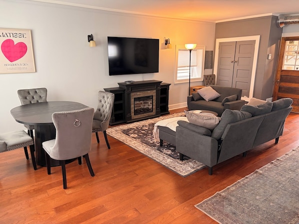 A living room with a fireplace, a flat-screen TV, a dining table with chairs, and a grey sofa.