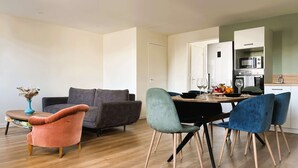 Una sala moderna con un sofá gris, un sillón rosa y sillas verde azulado. Una mesa de comedor con un jarrón de flores secas se encuentra en el centro.