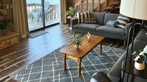 A spacious living room with a wooden staircase, a large window with a view of a lake, a grey sofa with 'welcome' pillows, and a wooden coffee table. - Sunset lake retreat: kayaks & NEW: boat rental! (Lake Ozark)