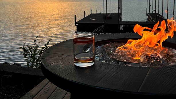 A glass of water on a deck overlooking a lake at sunset, with a fire pit and a covered dock in the background. - Sunset lake retreat: kayaks & NEW: boat rental! (Lake Ozark)