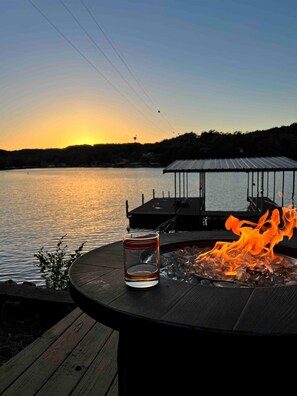 A glass of water on a deck overlooking a lake at sunset, with a fire pit and a covered dock in the background. - Sunset lake retreat: kayaks & NEW: boat rental! (Lake Ozark)