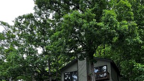 A two-story cabin with a wooden deck and a stone foundation near a body of water. - Sunset lake retreat: kayaks & NEW: boat rental! (Lake Ozark)