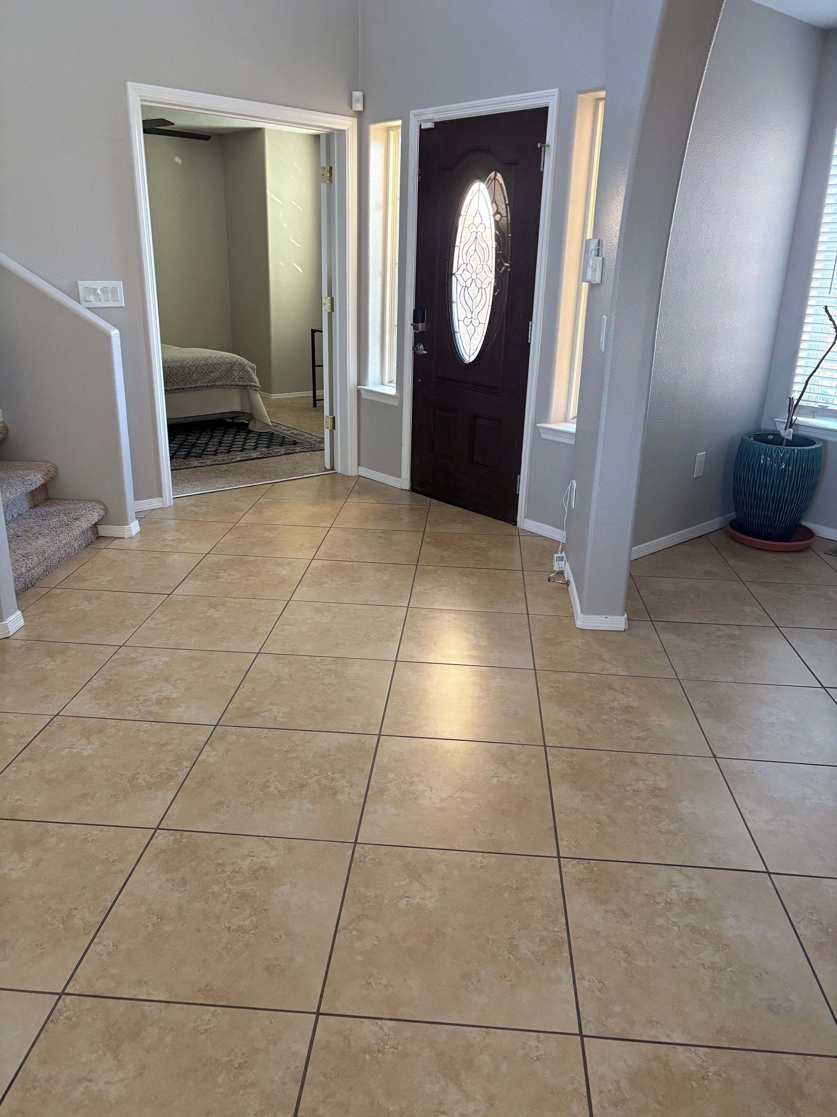 A spacious hallway with beige tiled flooring, a dark wooden door with a decorative glass window, and a staircase on the left.